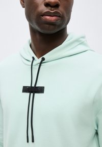 Κοντινό πλάνο ενός ατόμου που φοράει μια ανοιχτό πράσινη φούτερ με κουκούλα με μαύρα κορδόνια που περνούν μέσα από ένα μαύρο δερμάτινο έμπλαστρο στο στήθος.