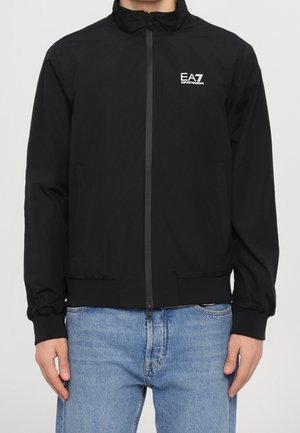 Homme portant une veste zippée noire EA7 Emporio Armani avec poignets et col côtelés, associée à un jean bleu clair.
