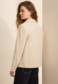 Beige bomullsjacka med krage, långa ärmar och knappmanchetter. Bakstycket har en ren, snedskuren fåll och en enkel design.