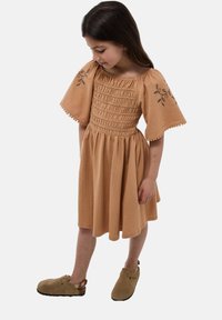 Robe couleur pêche en tissu texturé avec un buste smocké, des manches bouffantes et une broderie florale sur les manches. Portée avec des sabots marron.
