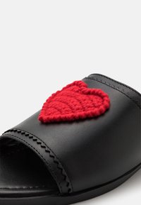 Black slide sandal with an open toe, featuring a textured red heart accent made of yarn on the upper, detailed stitching along the edges.