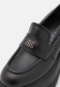 Mocassin en cuir noir avec un bout arrondi, présentant une plaque logo en argent bien visible sur une bande et une semelle en plateforme texturée.