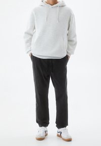 Felpa grigia chiara con cappuccio e tasca a marsupio, abbinata a pantaloni jogger neri scuri. Le scarpe presentano una base bianca con accenti neri e marroni.