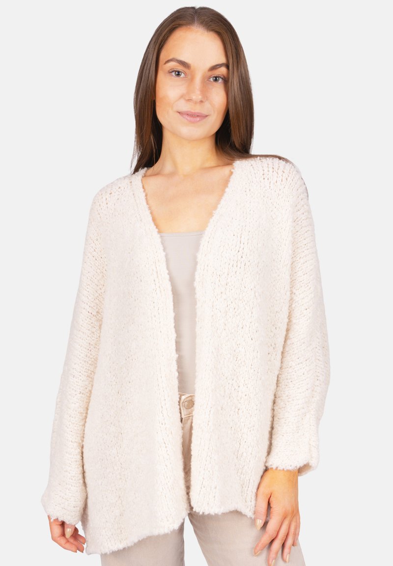 Frau in einer lockeren, weich texturierten, cremefarbenen offenen Strickjacke über einem beigen Oberteil und beigen Hosen, stehend vor einem schlichten Hintergrund.