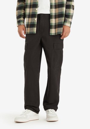 Schwarze Cargo-Hosen für Herren mit Seitentaschen, kombiniert mit weißen Sneakers und einem grün-beigen karierten Hemd mit Knöpfen, stehend vor weißem Hintergrund.