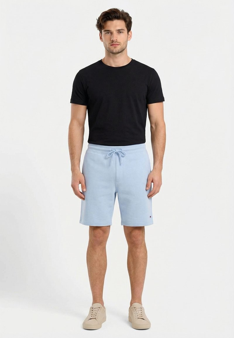 Jeune homme debout portant une chemise noire à manches courtes, un short bleu clair à cordon et des baskets beiges sur fond blanc.
