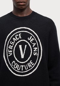 Črni pleteni pulover z velikim belim okroglem logotipom z napisom "VERSACE JEANS COUTURE" in osrednjo "V". Okrugla izrezana ovratnica in teksturirano blago.