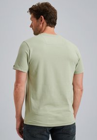 Svetlozelené bavlnené tričko s krátkymi rukávmi, okrúhlym výstrihom a malou obdĺžnikovou nášivkou na zadnej strane, skombinované s tmavými džínami.