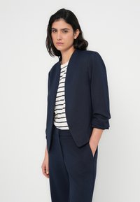 Μπλε σακάκι ναυτικού με άνετη εφαρμογή, που διαθέτει μανίκια τριών τετάρτων και λεία υφή. Συνδυασμένο με ριγέ μπλούζα, που εμφανίζει οριζόντιες γραμμές.