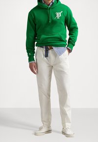 Grön bomulls-hoodie med en vit logotyp, kombinerad med krämfärgade byxor, kompletterad med en randig blåvit skjorta under.