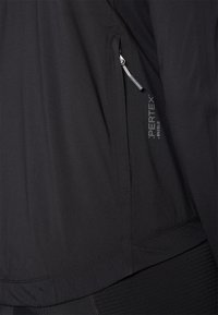 Veste noire en Pertex avec une finition texturée, dotée d'une poche zippée sur le côté et d'un logo discret, conçue pour une utilisation légère en extérieur.