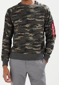 Felpa con motivo camouflage nei toni del verde scuro, marrone e nero, con polsini e orlo a costine. Presenta una tasca con zip sulla manica con un tirante rosso.