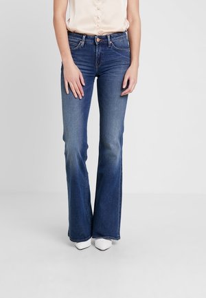 Femme portant un jean bleu taille haute évasé, un chemisier sans manches rose clair et des chaussures blanches pointues, debout les mains relâchées.