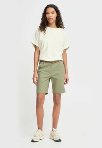 Kvinne med krøllete hår som har på seg en hvit kortermet skjorte, olivengrønne knelange shorts med et brunt belte, og hvite joggesko som står oppreist.