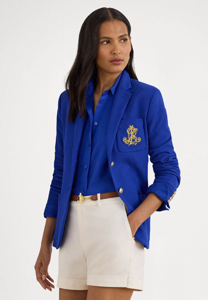 Kongelig blå blazer med gullknapper og brodert dekor på lommen, brukt over en matchende blå skjorte og beige shorts. Glatt tekstur.