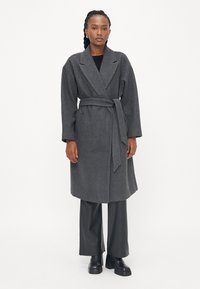 Cappotto avvolgente grigio in lana con vita alta e cintura, ampi risvolti e maniche larghe, abbinato a pantaloni neri in ecopelle e stivali neri chunky.