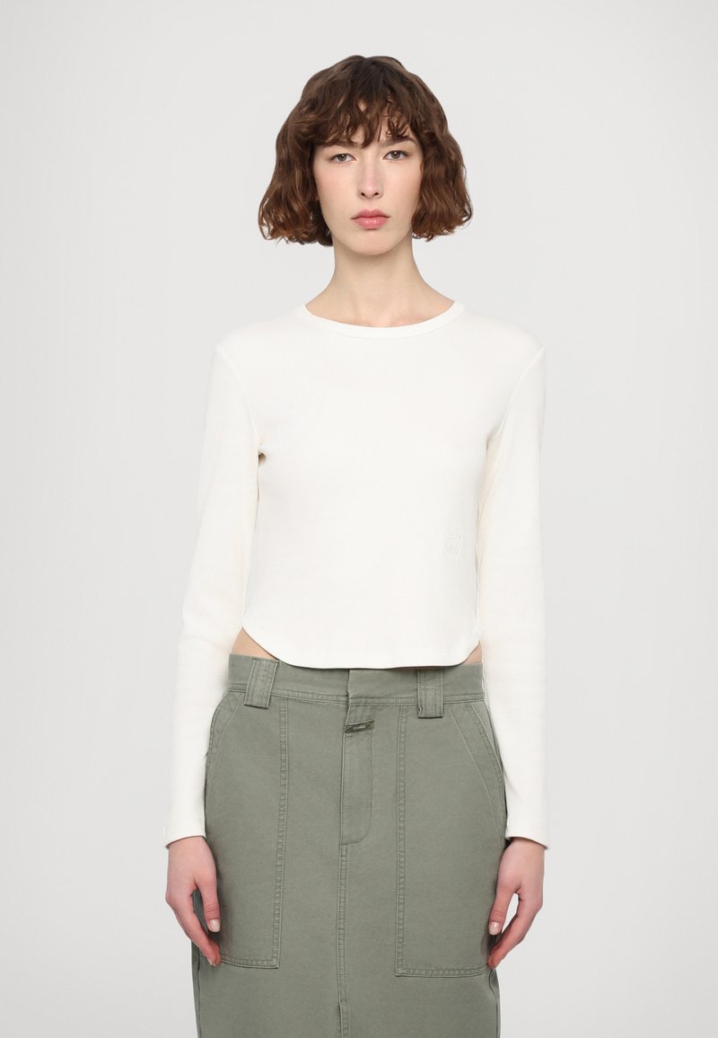 White long-sleeve cropped shirt with ribbed texture and a round neck, paired with a high-waisted olive green skirt featuring pockets.
