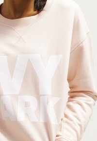 Sudadera rosa claro con cuello redondo. Presenta letras grandes en blanco que dicen "IVY PARK" en la parte delantera. La tela parece suave y tiene puños acanalados.