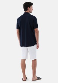 Marineblaues kurzärmliges Hemd mit Kragen, kombiniert mit weißen Shorts. Das Hemd hat eine glatte Textur und einen abgerundeten Saum. Die Schuhe sind schwarze Sandalen.