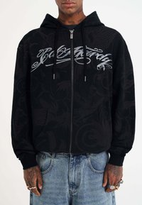 Black zip-up hoodie featuring a textured design, front pockets, and silver graphic lettering. Material appears soft with ribbed cuffs.