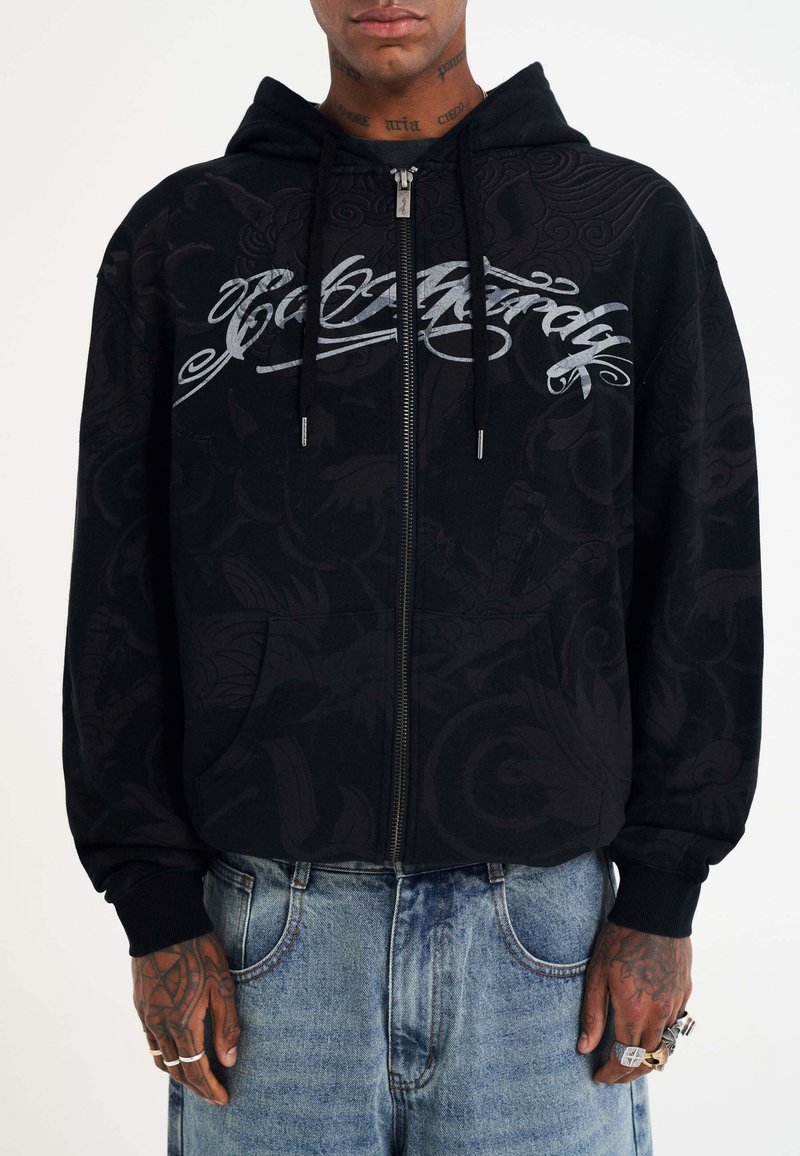 Schwarze Zip-Up-Hoodie mit strukturiertem Design, frontalen Taschen und silberner Grafikbeschriftung. Das Material sieht weich aus und hat gerippte Bündchen.