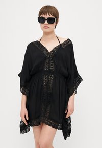 Kaftán negro con intrincado borde de encaje, con un profundo escote en V, mangas cortas y un corte holgado, combinado con unas grandes gafas de sol negras.