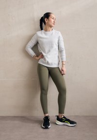 Grå hettegenser, olivengrønne leggings og svarte joggesko med gule detaljer. Bakgrunnen er en lys, teksturert vegg.