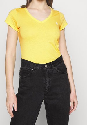 Femme portant un t-shirt jaune à manches courtes et col en V, rentré dans un jean noir taille haute, sur un fond uni.