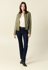 Giacca in pelle verde oliva con chiusura a scatto, blusa crema sotto, e jeans a zampa d'elefante blu scuro, abbinati a stivali neri alla caviglia.