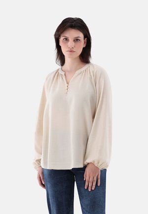 Helle beigen Bluse mit langen Ärmeln, gerafftem Ausschnitt und einer Knopfleiste mit drei Knöpfen. Geschmeidige Textur und lockere Passform. Kombiniert mit blauen Jeans.