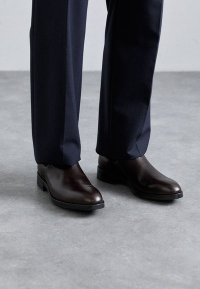 Zapatos de vestir de cuero marrón con un diseño elegante, punta redonda y tacón bajo, combinados con pantalones oscuros a medida sobre una superficie gris texturizada.