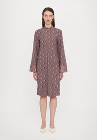 Langärmliges, mauvefarbenes Kleid mit geometrischem Muster, das dunkelgrüne Rauten zeigt. Hoher Ausschnitt und weicher Stoff, der bis zur Mitte der Waden reicht.