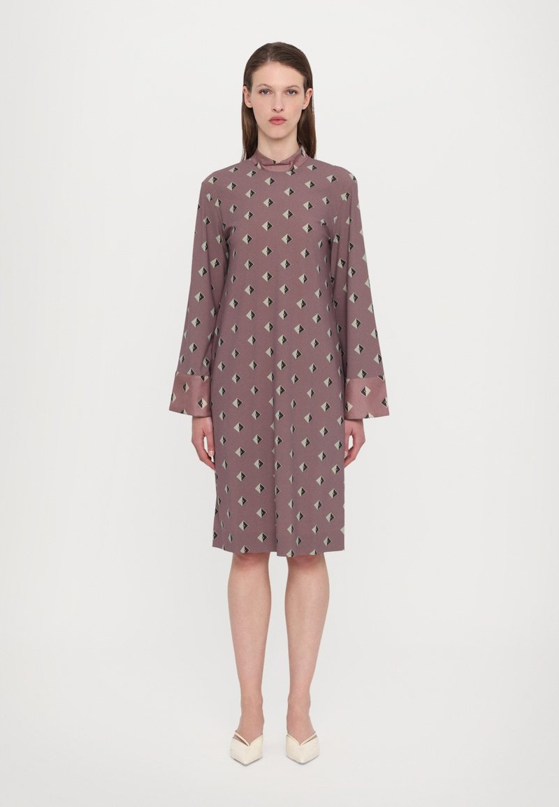 Langärmliges, mauvefarbenes Kleid mit geometrischem Muster, das dunkelgrüne Rauten zeigt. Hoher Ausschnitt und weicher Stoff, der bis zur Mitte der Waden reicht.