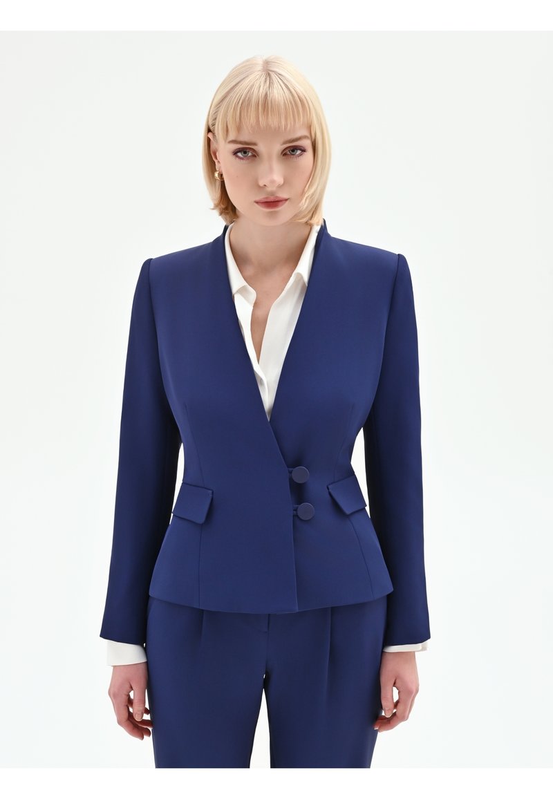 Blonde vrouw met gouden oorringen en een op maat gemaakt marineblauw pak met dubbele knopen over een witte blouse, staand tegen een witte achtergrond.