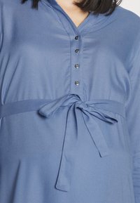 Camicia da uomo in denim blu con scollatura a V, maniche lunghe e cintura con lacci in vita. Presenta bottoni lucidi e un tessuto morbido e liscio.