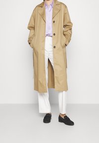 Personne portant un trench-coat beige sur une chemise lavande avec un pantalon blanc et des mocassins noirs, debout devant un fond uni clair.