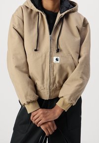 Beige capuchonjack gemaakt van een katoenmix, met een voorkoord, zwarte accenten en een logo-patch. Geribbelde manchetten, losse pasvorm, zachte textuur.