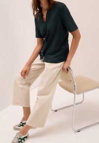 Femme assise sur une chaise beige portant un chemisier vert foncé à manches courtes, un pantalon large crème et des baskets beiges avec des rayures vertes.