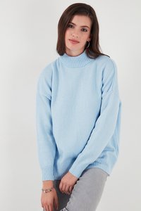 Nuori nainen, jolla on ruskeat hiukset ja joka pukeutunut vaaleansiniseen neuleeseen ja harmaisiin housuihin, poseeraa yksinkertaisen valkoisen taustan edessä.