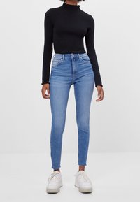 Top corto negro de manga larga combinado con jeans skinny de tiro alto en azul, con un lavado desgastado y dobladillo deshilachado. Zapatillas blancas completan el conjunto.