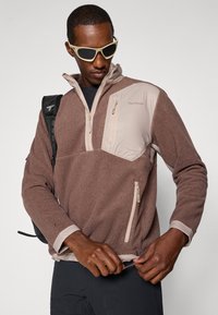 Pull en polaire marron avec un col montant, accents beige clair, poches zippées et cordon de serrage à l'ourlet, présentant une finition texturée.
