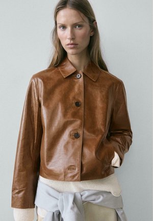 Mladá žena s dlouhými vlasy, která má na sobě hnědou koženou zapínanou bundu, krémový pletený svetr a světle šedý oděv přivázaný v pase.