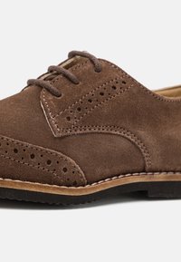 Ruskea mokkanahkainen nyörikenkä, jossa on rei'itetyt yksityiskohdat, kontrastisauma ja teksturoitu musta kumipohja. Klassinen oxford-malli.