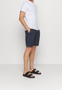 Camiseta blanca con lunares azul marino y un logo amarillo, combinada con pantalones cortos azul marino y sandalias negras tipo slide con dos correas y hebillas.