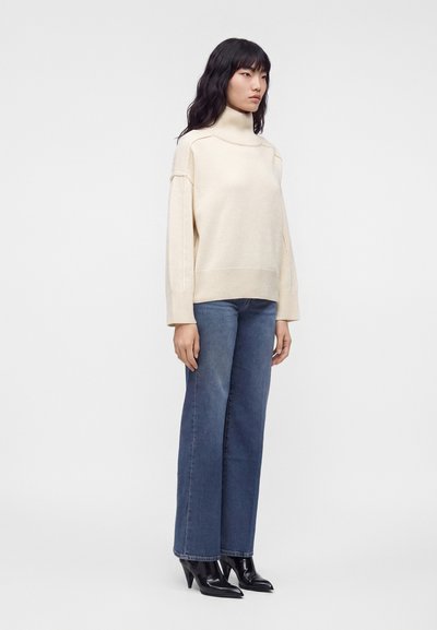 Femme portant un pull à col roulé crème, un jean bleu droit et des bottines noires à talons, debout devant un fond clair uni.