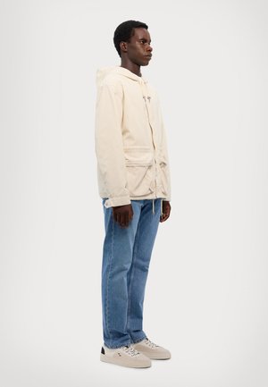 Veste beige clair avec une capuche, taille à cordon de serrage et poches avant, associée à un jean bleu et des baskets beiges. Design simple, style décontracté.