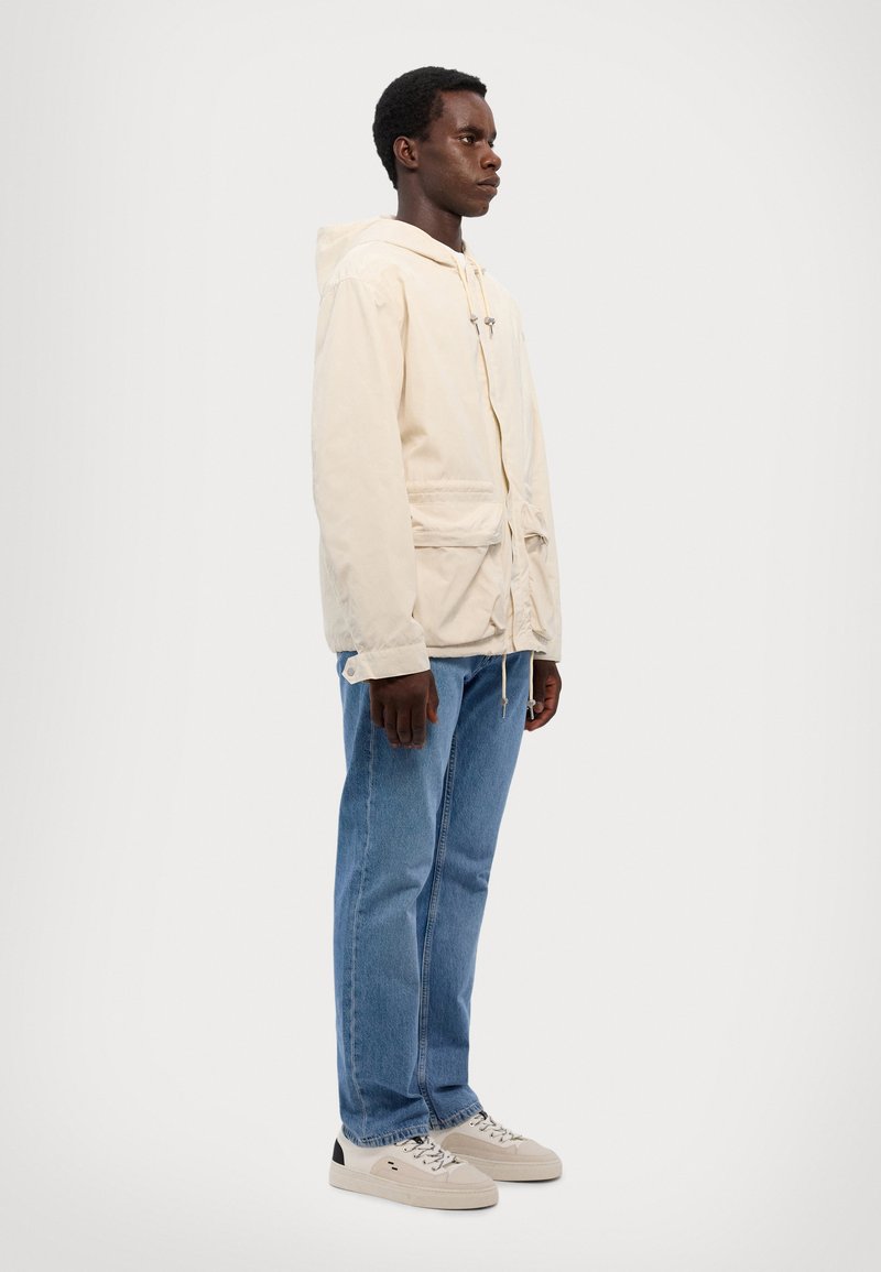 Svetlo bež jakna s kapuco, s vrvico v pasu in sprednjimi žepi, v kombinaciji z modrimi kavbojkami in bež športniki. Preprost dizajn, sproščen stil.