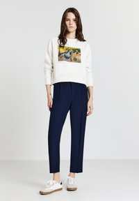 Giovane donna che indossa una felpa bianca con stampa di un dipinto di Paul Gauguin, pantaloni tapered blu navy e sneakers bianche, in piedi contro uno sfondo neutro.