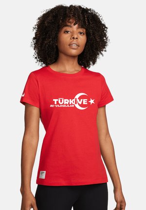 Femme aux cheveux bouclés portant un t-shirt rouge avec un croissant et une étoile turcs blancs et le mot "Türkive" sur un fond uni.