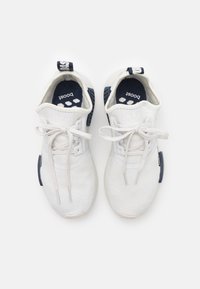 Vita idrottsskor med stickad ovandel, vita snören och marinblå accent på sulan. Har ett vadderat inre och dragtab.